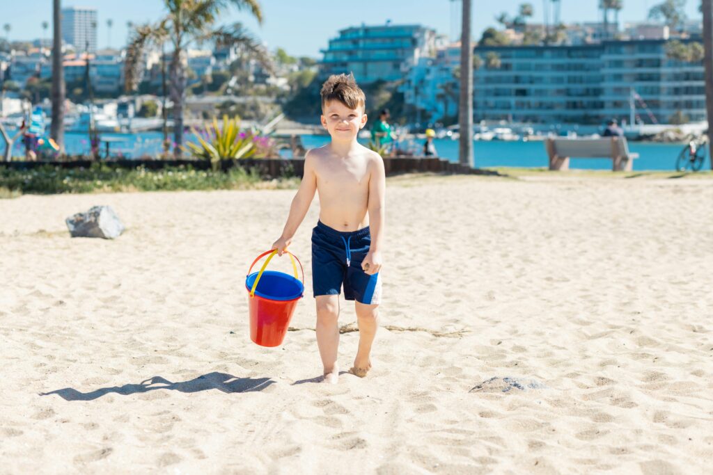 A joyful child carrying a red bucket on a sandy beach during a sunny day by the ocean.