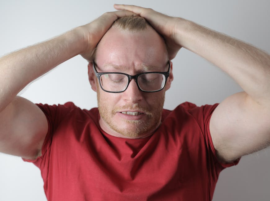 Irritated bearded man in red tee shirt and eyewear straightening hair back with closed eyes while standing against gray background
